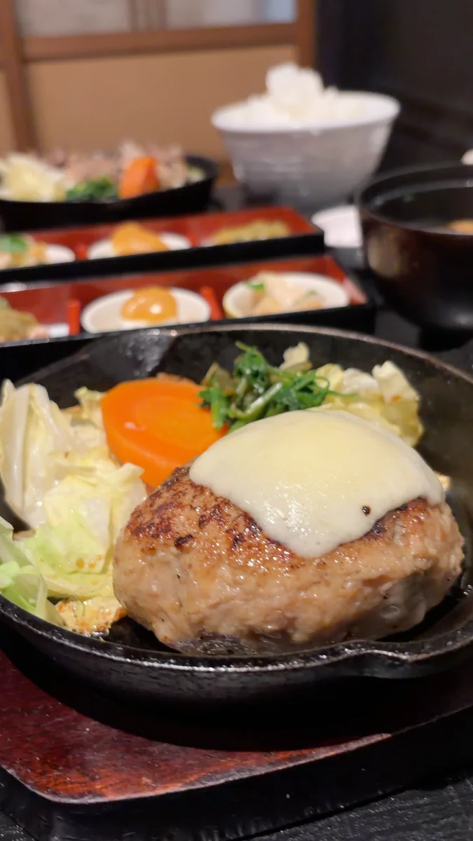 2人分の膳。上がハンバーグ定食、下が炭焼き定食で、どちらにも三種盛・汁・香の物・ごはんが付きます(左)/ハンバーグの上にチーズ。鉄のスキレットのまま出てきます(右)